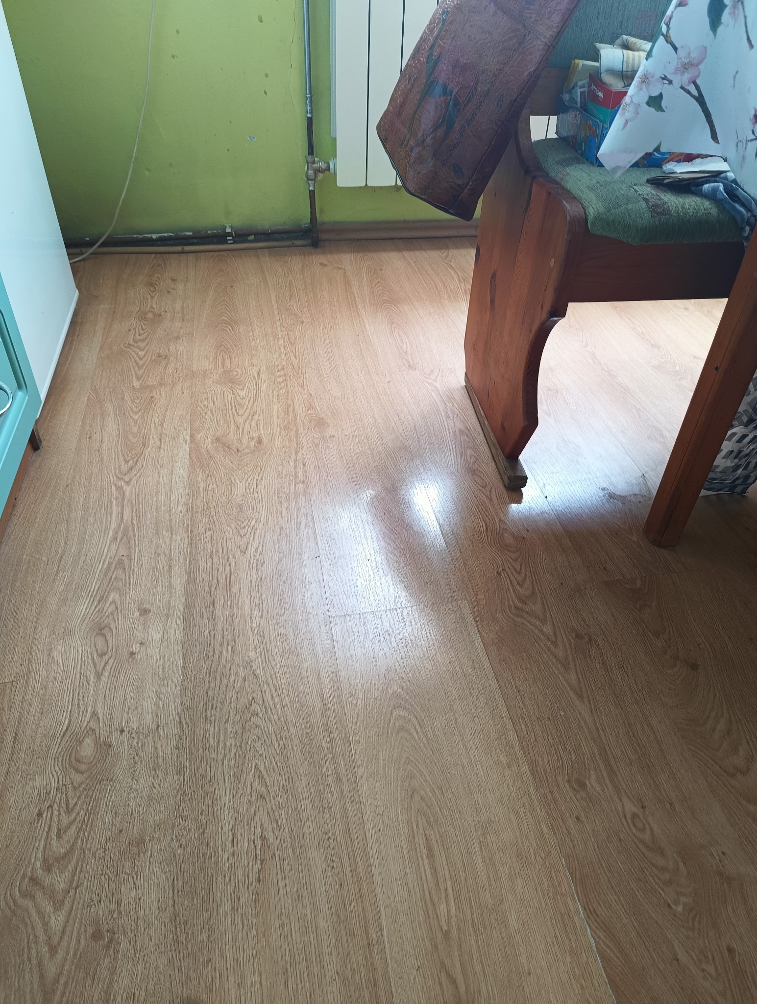 Jasne panele podłogowe z widoczną strukturą drewna, fragment ściany w kolorze zielonym, oraz fragment drewnianego krzesła w tle.