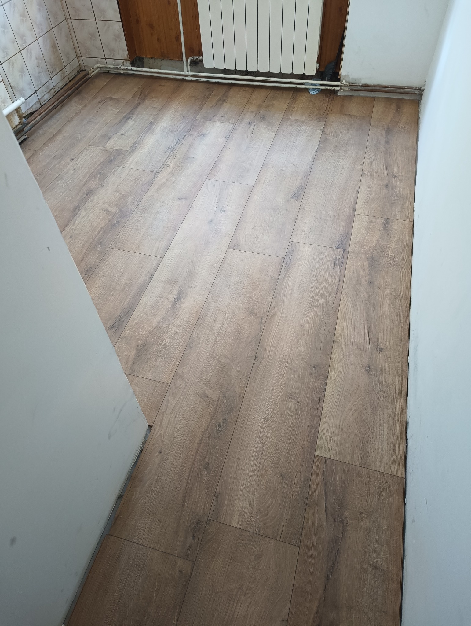 Ukończona podłoga z paneli w odcieniach brązu, widok z góry, z fragmentem białej ściany i grzejnika. Widoczne detale łączeń paneli i tekstury drewna.