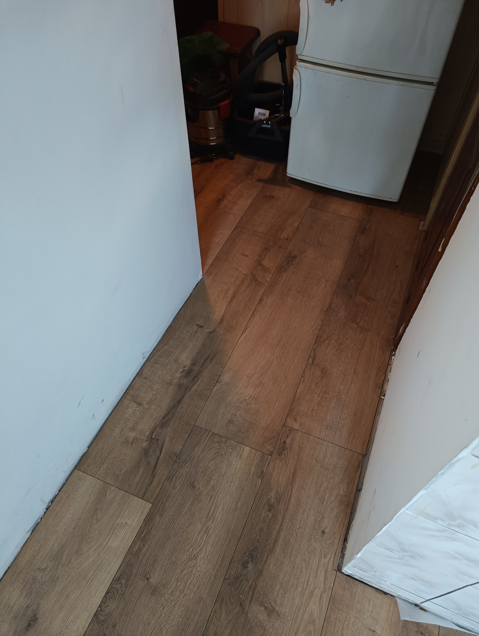 Ułożone panele podłogowe w wąskim pomieszczeniu, widoczne białe ściany i fragmenty sprzętów AGD. Układanie paneli z wykończeniem przy ścianie.