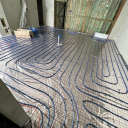 Vasyl Mykolaichuk - Instalacja ogrzewania podłogowego w Grójcu. Widoczne niebieskie rury ułożone na izolacji z siatką, przygotowane do zalania wylewką. W tle surowe ściany i drewniane drzwi.