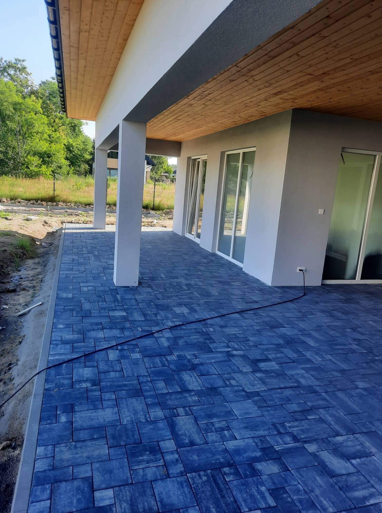 Niebieska kostka brukowa na tarasie domu z drewnianym podbitką. Widoczne słupy podtrzymujące zadaszenie i częściowo przygotowany teren pod dalsze prace.