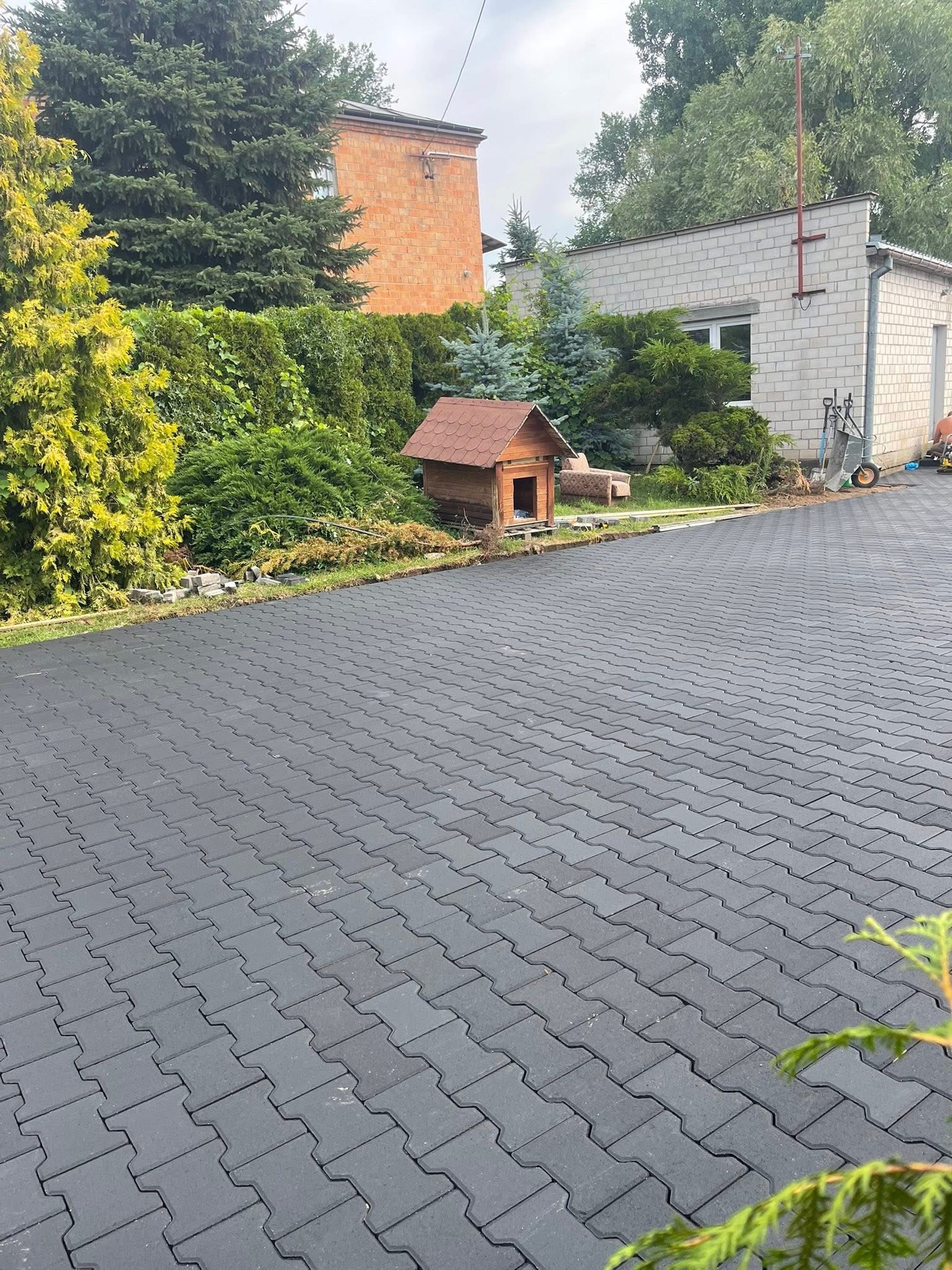 Ułożona, szara kostka brukowa na podjeździe, z widocznym fragmentem ogrodu i budynkiem w tle. Charakterystyczny wzór kostki, regularny układ.