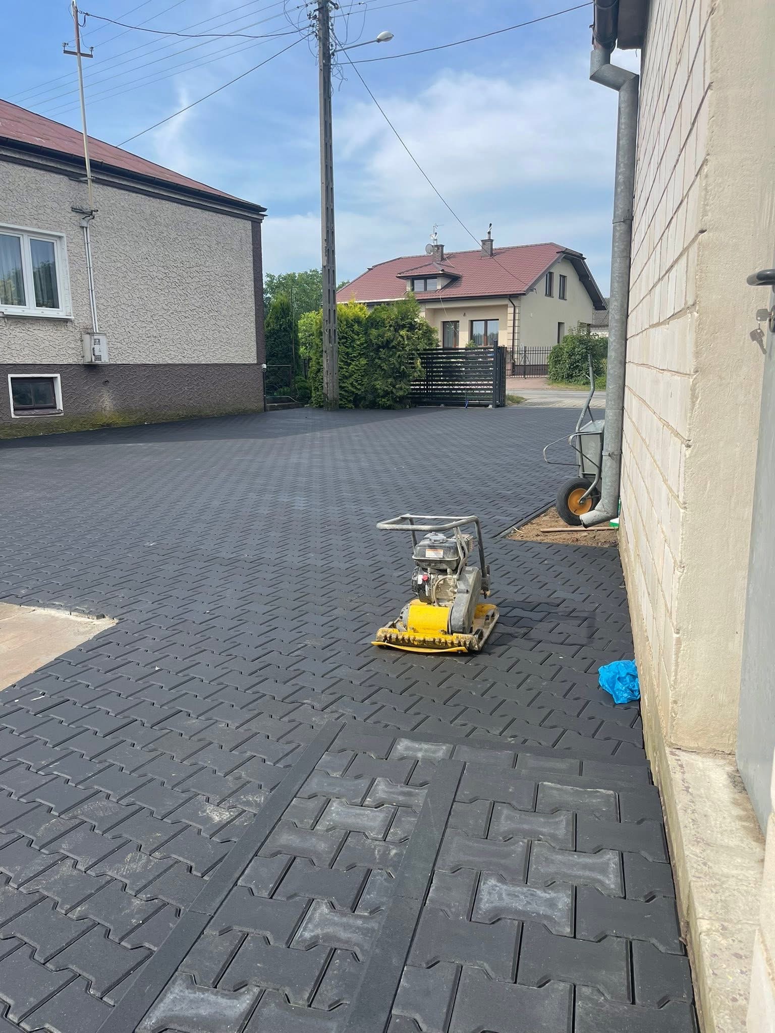 Ułożona, grafitowa kostka brukowa typu I na podjeździe, z żółtą zagęszczarką w kadrze. W tle budynki mieszkalne i błękitne niebo.