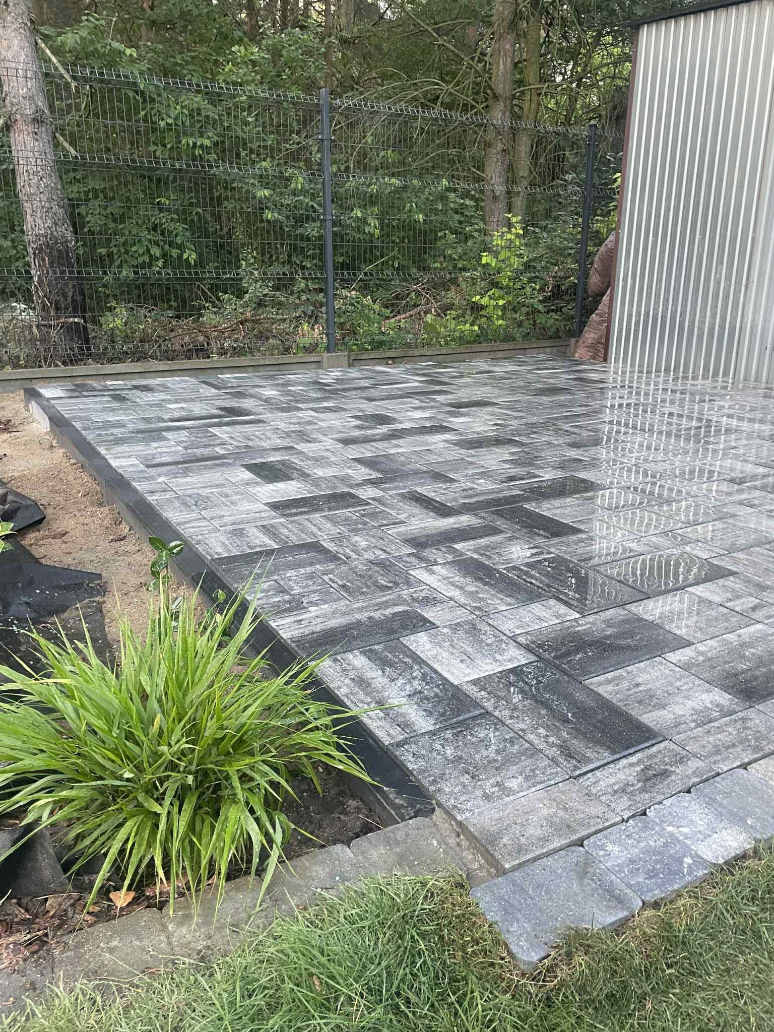 Ułożona, szara kostka brukowa w geometryczny wzór, obramowana zielenią trawy i roślin. W tle metalowa konstrukcja i drzewa.