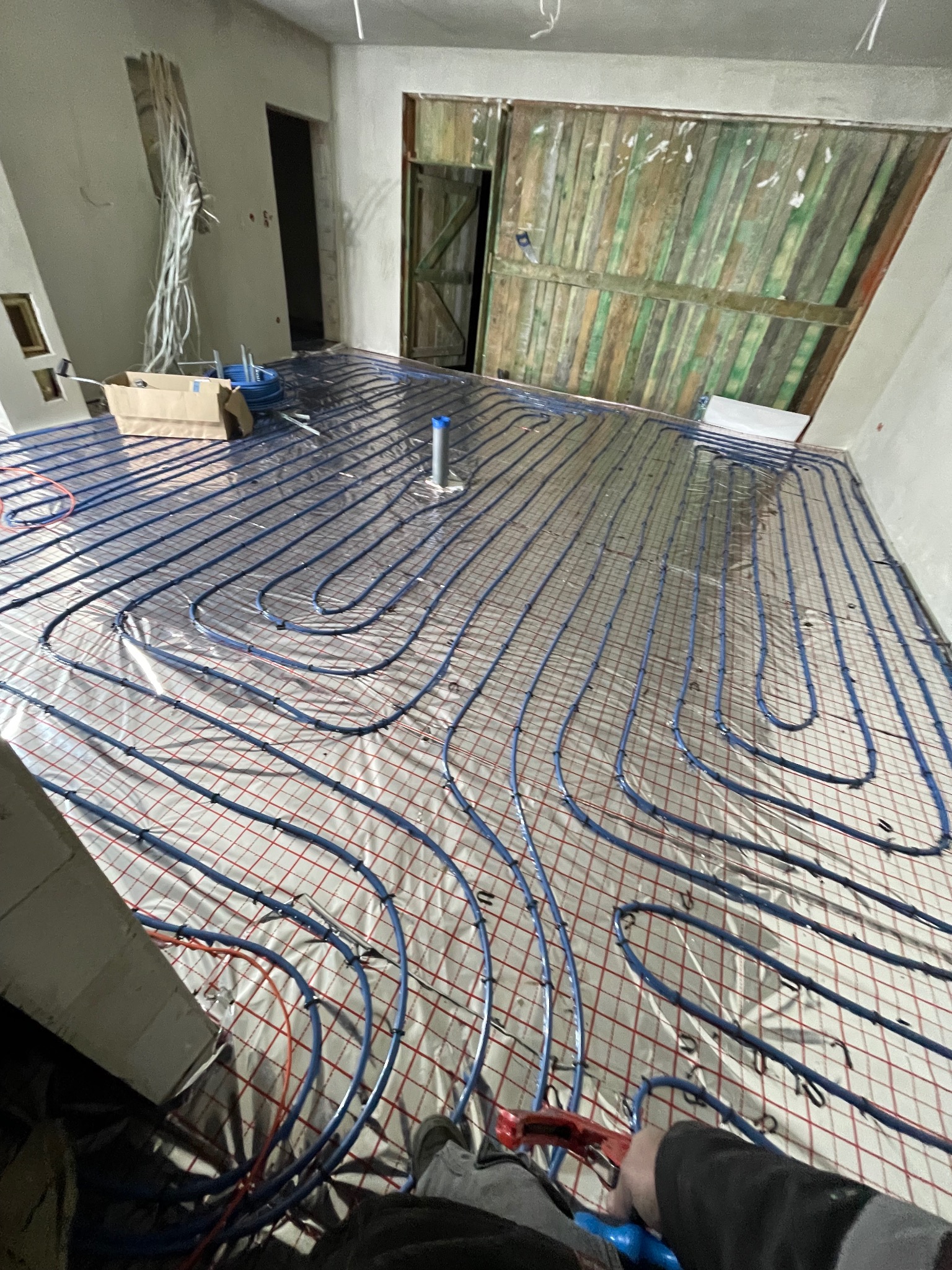 Instalacja ogrzewania podłogowego w Grójcu. Widoczne niebieskie rury ułożone na izolacji z siatką, przygotowane do zalania wylewką. W tle surowe ściany i drewniane drzwi.