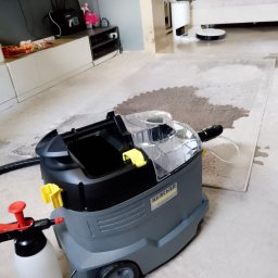 CZYSTA ROBOTA-EKIPA NA BŁYSK - Przemysłowy odkurzacz piorący Karcher na tle częściowo wyczyszczonego, jasnego dywanu w pokoju. Widoczny spryskiwacz i wąż ssący.