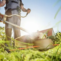 Mężczyzna kosi trawę kosiarką spalinową, widok z dołu na ostrze tnące, w tle dom i błękitne niebo. Promienie słoneczne.