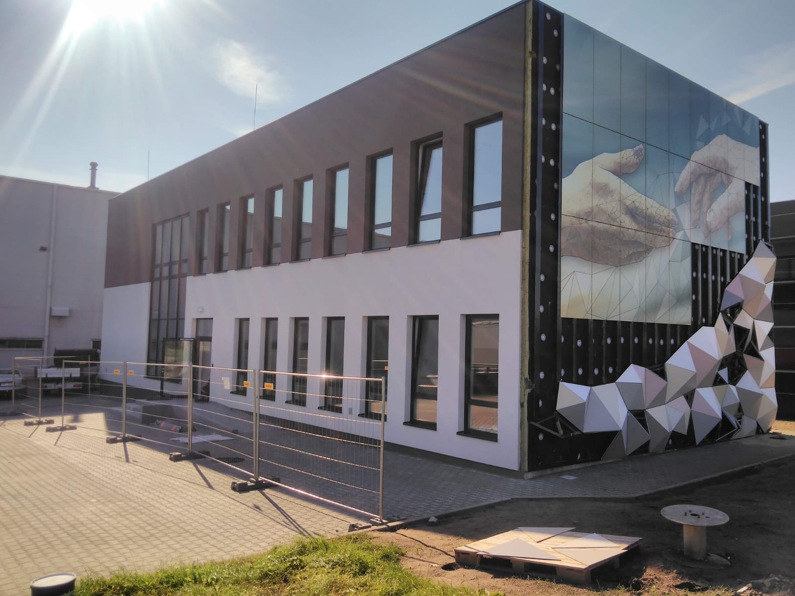 Elewacja budynku z motywem dłoni i geometrycznymi elementami dekoracyjnymi, widoczne rusztowania i teren budowy. Częściowo ukończona fasada.