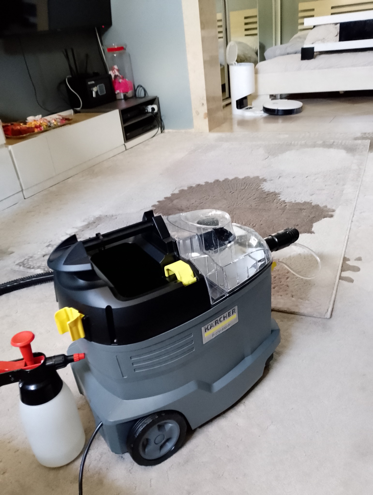 Przemysłowy odkurzacz piorący Karcher na tle częściowo wyczyszczonego, jasnego dywanu w pokoju. Widoczny spryskiwacz i wąż ssący.