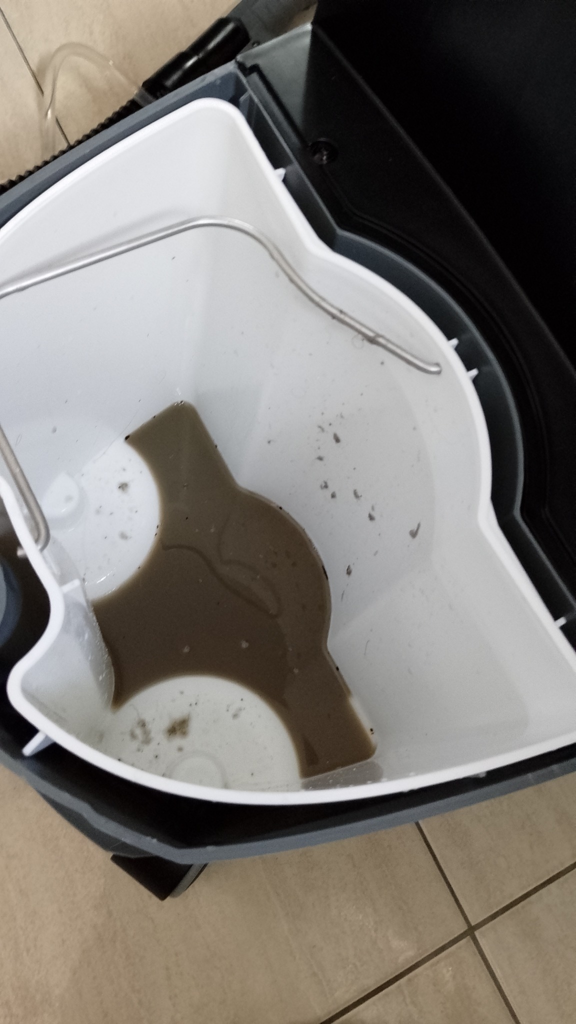 Zbiornik odkurzacza po praniu tapicerki, wypełniony brudną wodą i osadami. Widoczne drobinki zanieczyszczeń. Ujęcie z góry, na beżowym tle płytek podłogowych.