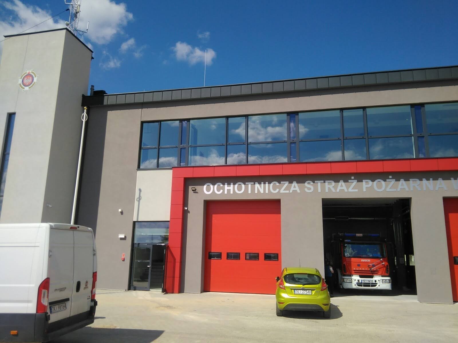 Budynek Ochotniczej Straży Pożarnej z czerwonymi bramami garażowymi, zaparkowanym autem i wozem strażackim wewnątrz. Błękitne niebo z chmurami w tle.