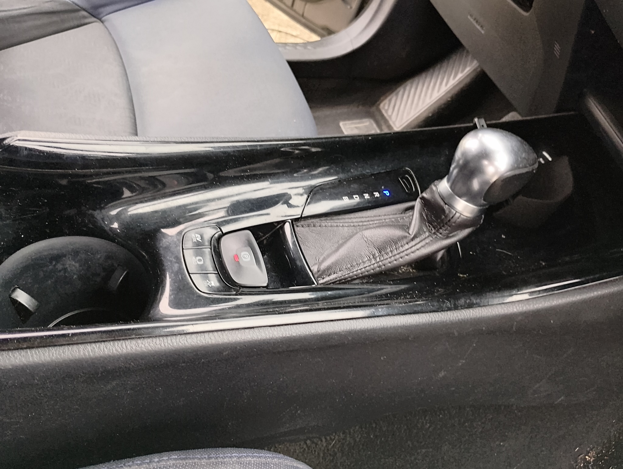 Błyszcząca, czarna konsola środkowa samochodu z dźwignią zmiany biegów i przyciskami funkcyjnymi, widoczne ślady użytkowania.