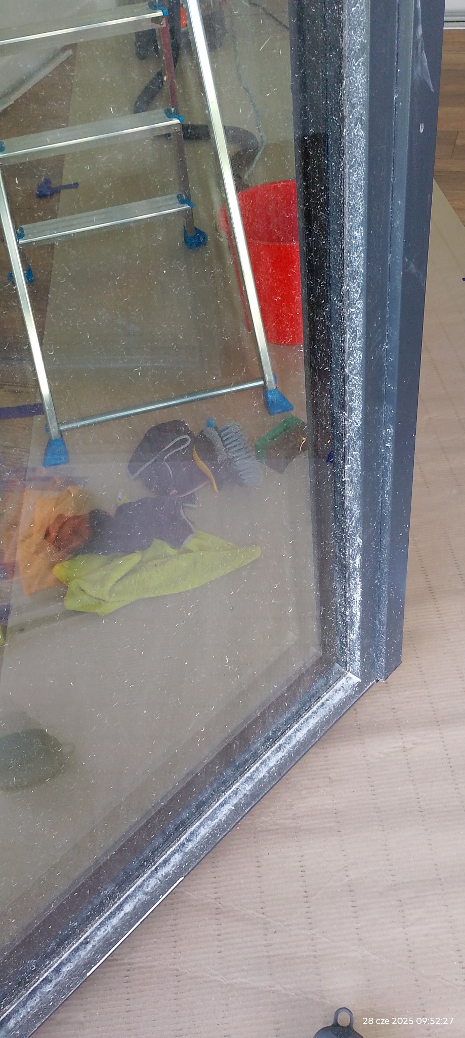 Brudna szyba okienna z widocznymi smugami, drabina, szczotki i wiadro w tle. Szyba w ciemnej ramie, widoczna data w rogu.