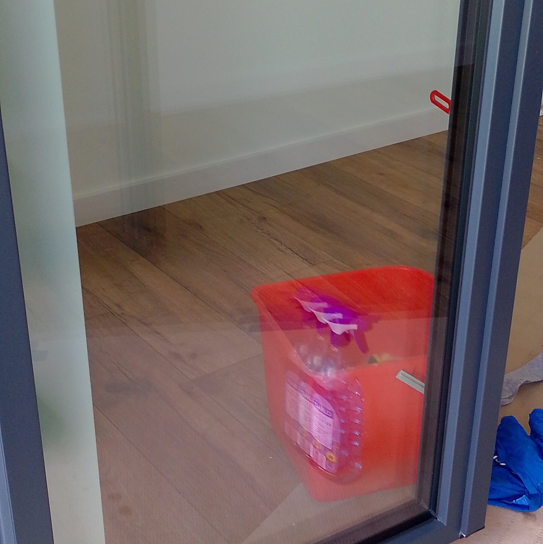 Wnętrze z widocznym oknem, na podłodze stoi czerwone wiadro z płynem do mycia okien i niebieskie rękawice. Świeżo umyte okno odbija elementy wnętrza.