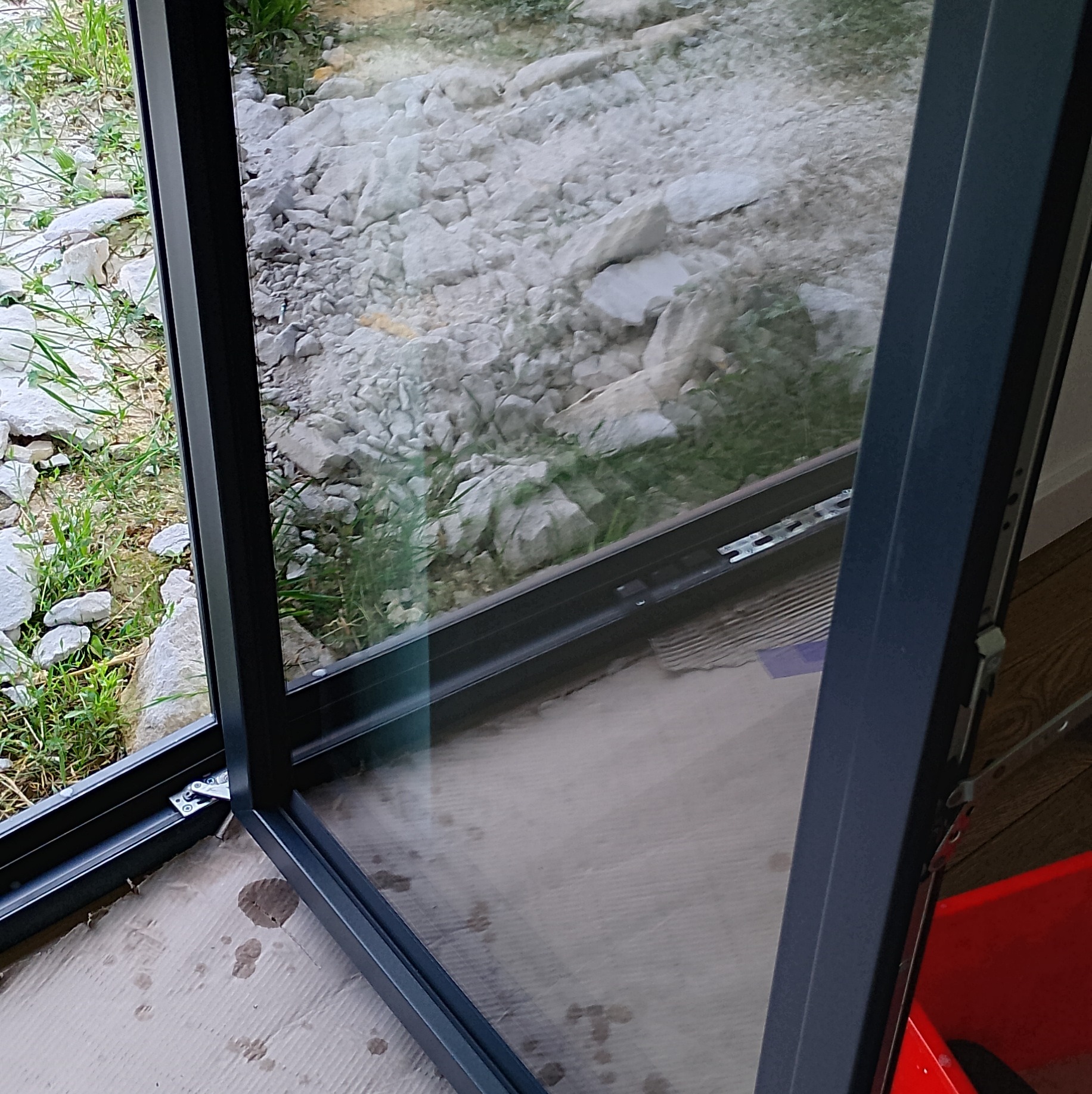 Zamontowane okno z widokiem na kamieniste podłoże i trawę. Nowoczesna rama okienna w kolorze antracytowym, widoczne detale montażowe.