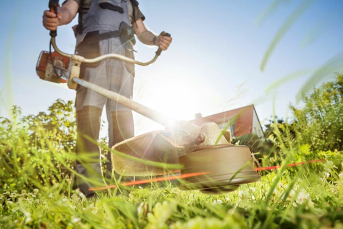 Mężczyzna kosi trawę kosiarką spalinową, widok z dołu na ostrze tnące, w tle dom i błękitne niebo. Promienie słoneczne.