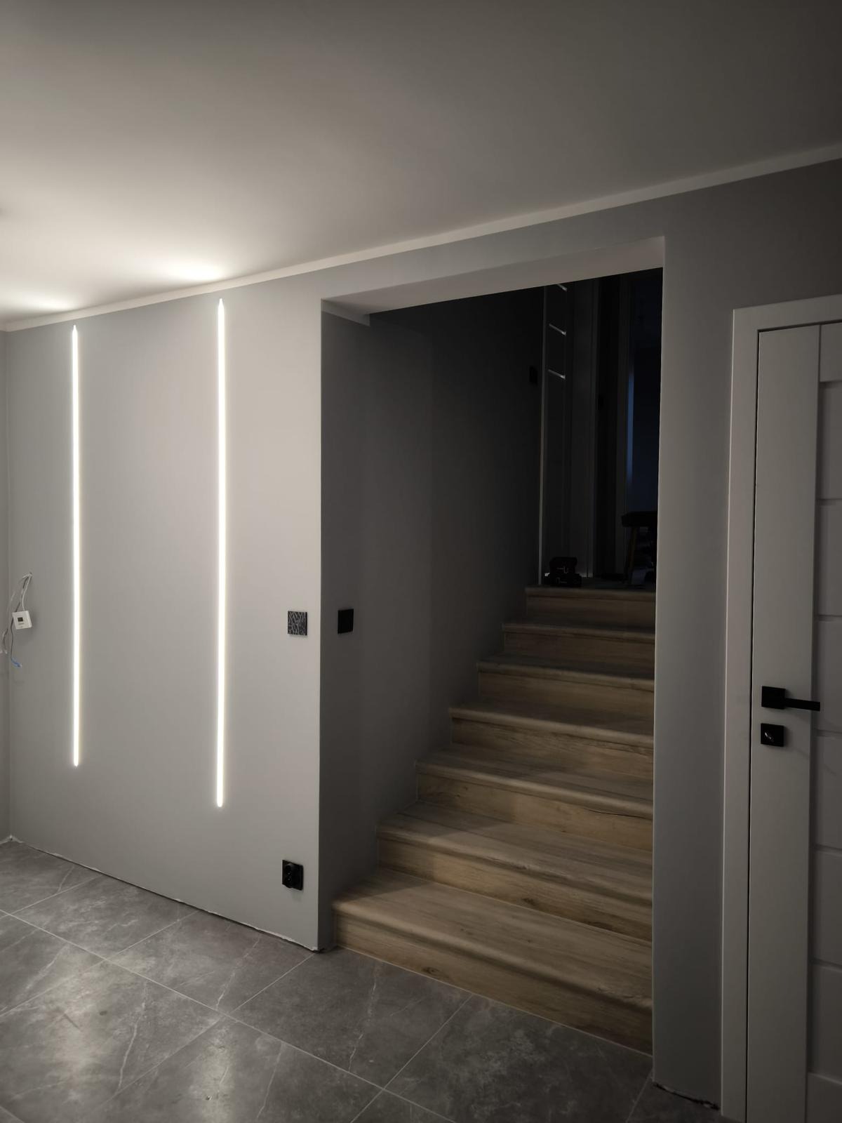 Nowoczesne wnętrze: szare ściany z pionowymi paskami LED, drewniane schody i grafitowa podłoga. Minimalistyczny design i subtelne oświetlenie.