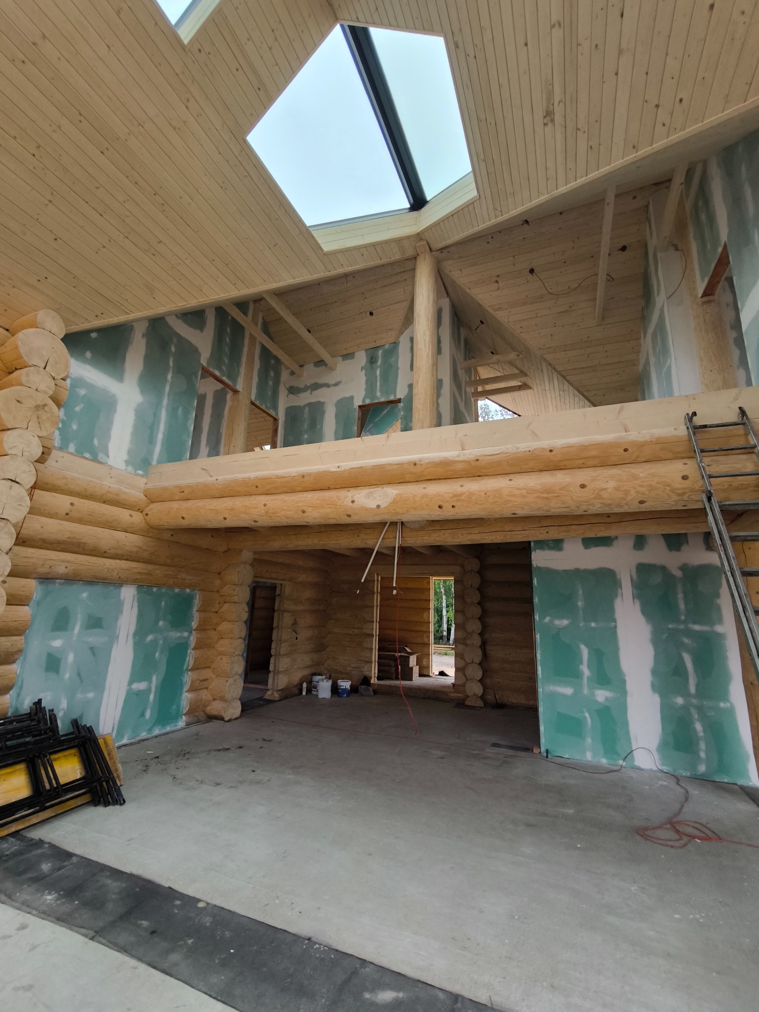 Wnętrze domu z bali w trakcie prac wykończeniowych. Widoczne płyty kartonowo-gipsowe na ścianach, drewniany strop z oknem dachowym i drabina.