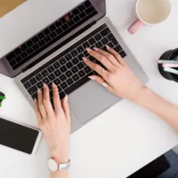 Kobieta pisze na laptopie na białym biurku z telefonem, rośliną i przyborami. Widok z góry, skupienie na dłoniach z biżuterią i manicure.