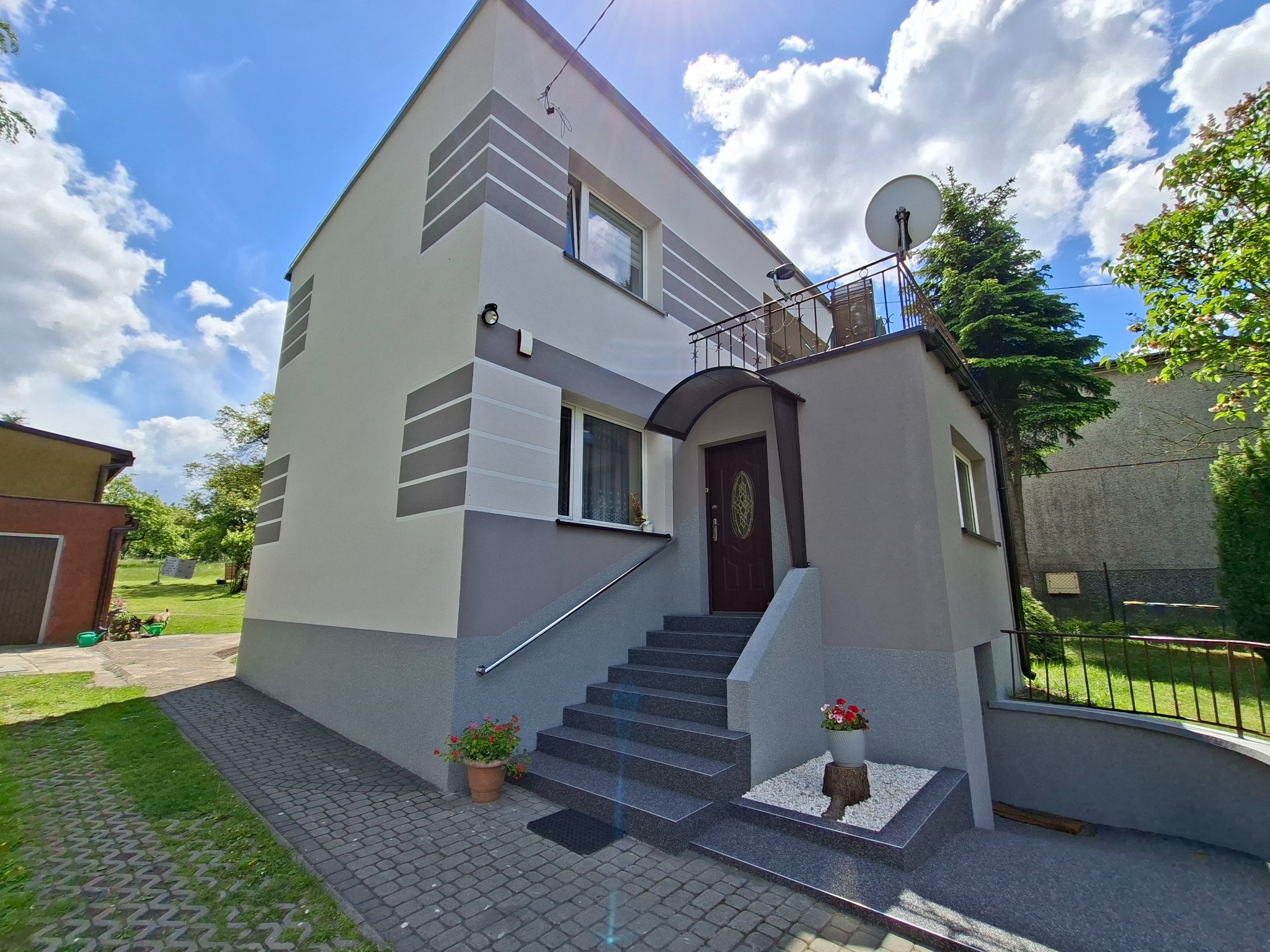 Dwukondygnacyjny dom z elewacją w odcieniach szarości, z ciemnymi pasami i schodami wejściowymi. Widoczne kwiaty w donicach i satelita na balkonie.