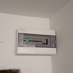 SMART-TECH Instalacje Elektryczne Patryk Jurdziński - Zamknięta, biała skrzynka elektryczna Elektro-Plast na białej ścianie, z widocznymi bezpiecznikami i opisami obwodów 1-10. Świeci się zielona dioda.
