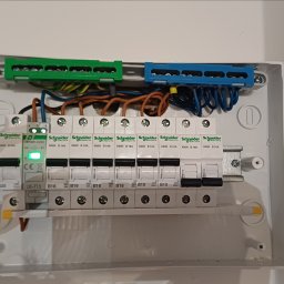 SMART-TECH Instalacje Elektryczne Patryk Jurdziński - Rozdzielnica elektryczna Schneider Electric z bezpiecznikami B16 i B10, z zielonym i niebieskim panelem zaciskowym, widoczne przewody.