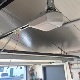 Zamontowany napęd do bramy garażowej z czerwoną linką awaryjnego otwierania, widok z wnętrza garażu, na zewnątrz widoczny fragment domu.