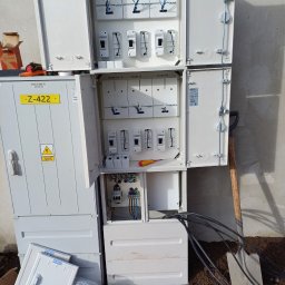 SMART-TECH Instalacje Elektryczne Patryk Jurdziński - Otwarta, biała szafa elektryczna zewnętrzna z bezpiecznikami i przewodami, z żółtą tabliczką Z-422. Obok leżą pokrywy i narzędzia, a w tle widać szpadel.