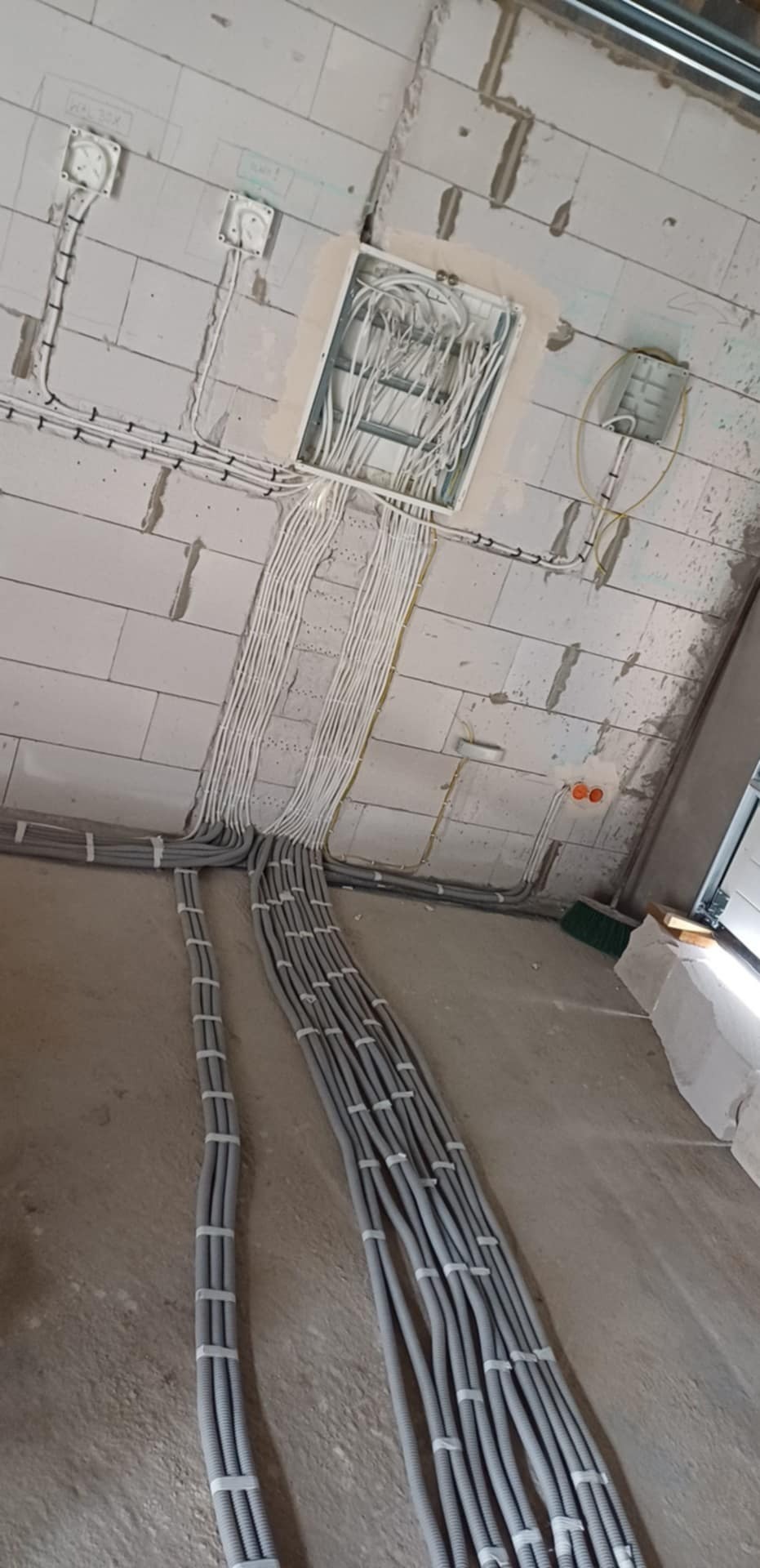 Szara, karbowana rura instalacyjna ułożona równolegle na podłodze, prowadząca do białej skrzynki elektrycznej na ścianie z bloczków. Widoczne kable.