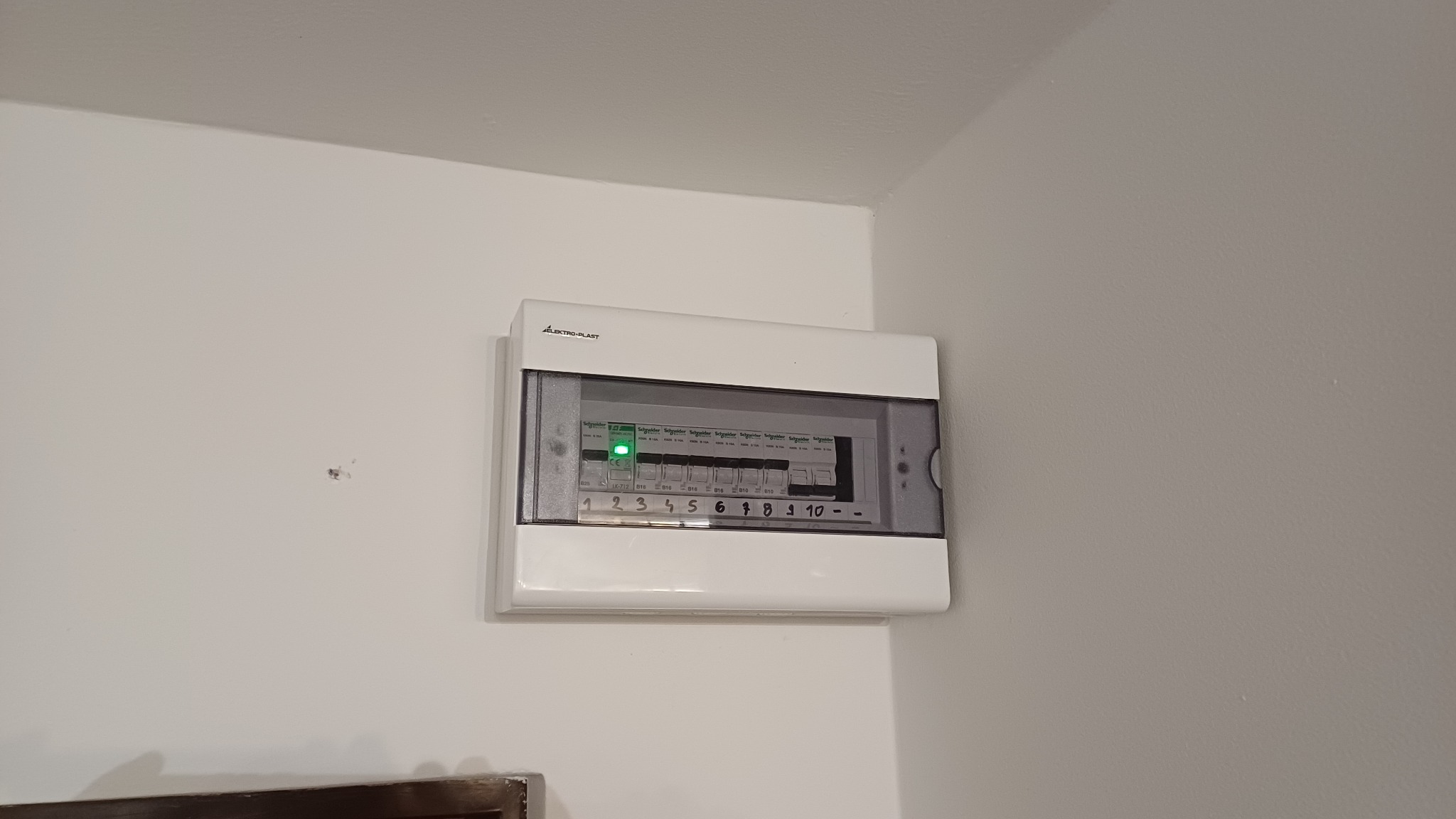 Zamknięta, biała skrzynka elektryczna Elektro-Plast na białej ścianie, z widocznymi bezpiecznikami i opisami obwodów 1-10. Świeci się zielona dioda.