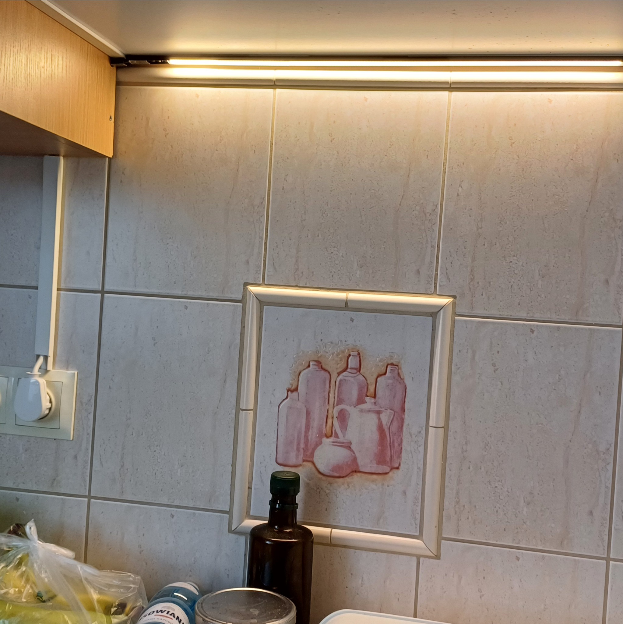 Fragment kuchni z oświetleniem podszafkowym LED, kremowymi płytkami i gniazdkiem elektrycznym. Widoczny także obrazek na ścianie i butelka oleju.