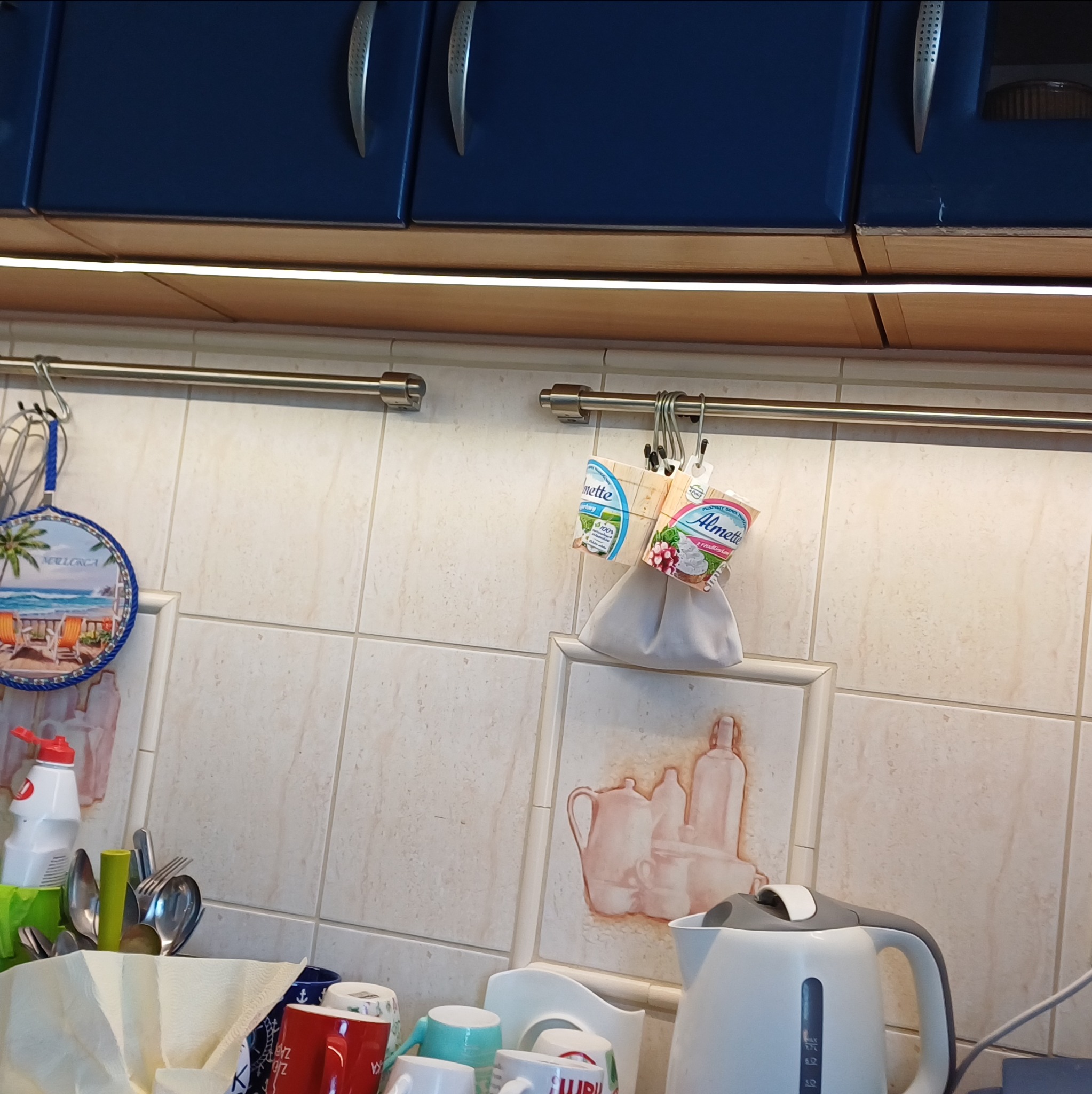 Kuchnia z niebieskimi szafkami, oświetleniem LED pod szafkami, relingiem z akcesoriami i płytkami w tle. Widoczny czajnik i naczynia.