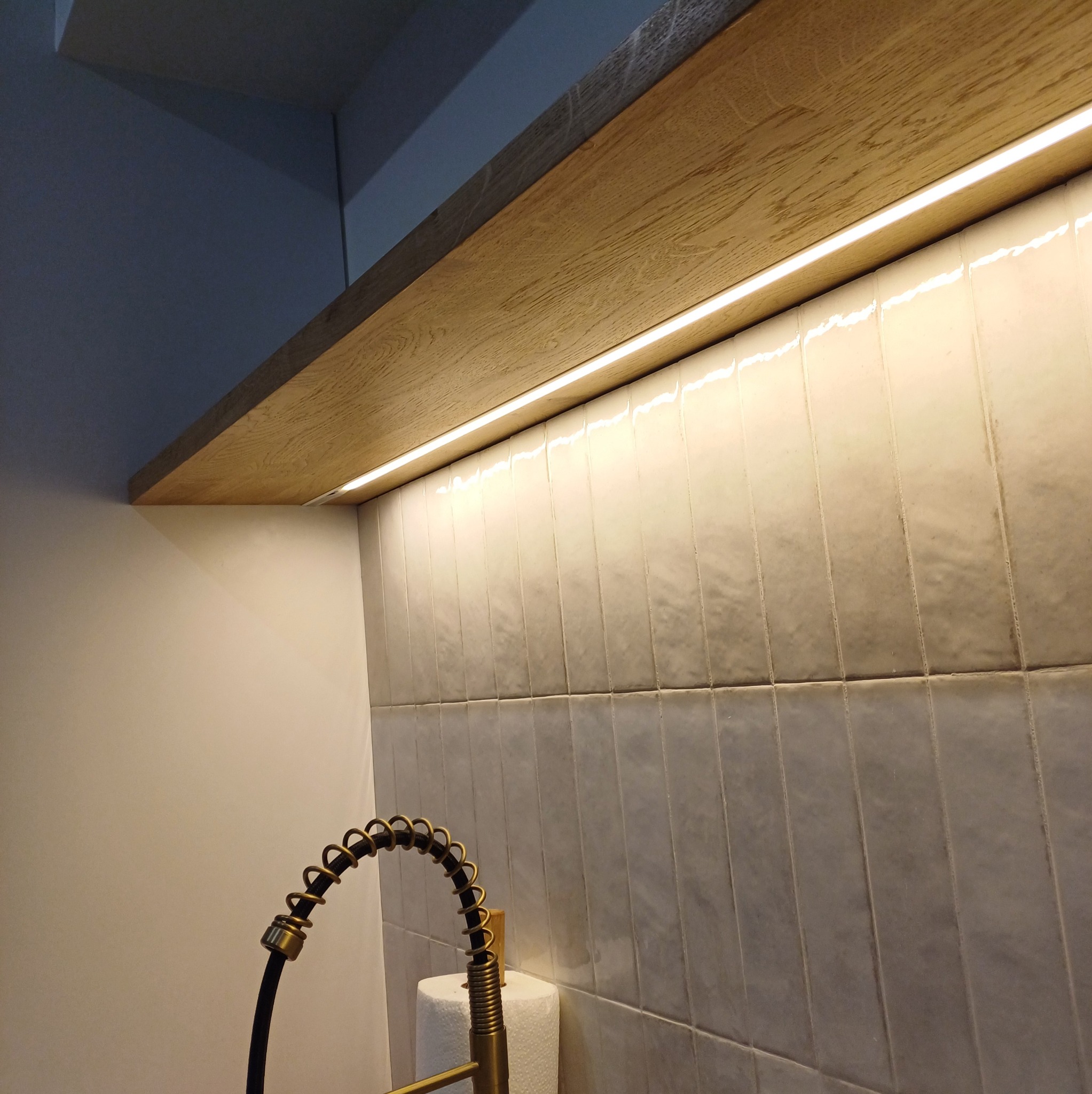 Oświetlenie LED zamontowane pod drewnianą półką, oświetlające ceramiczne płytki na ścianie nad zlewem z baterią w kolorze starego złota. Ciepłe światło.