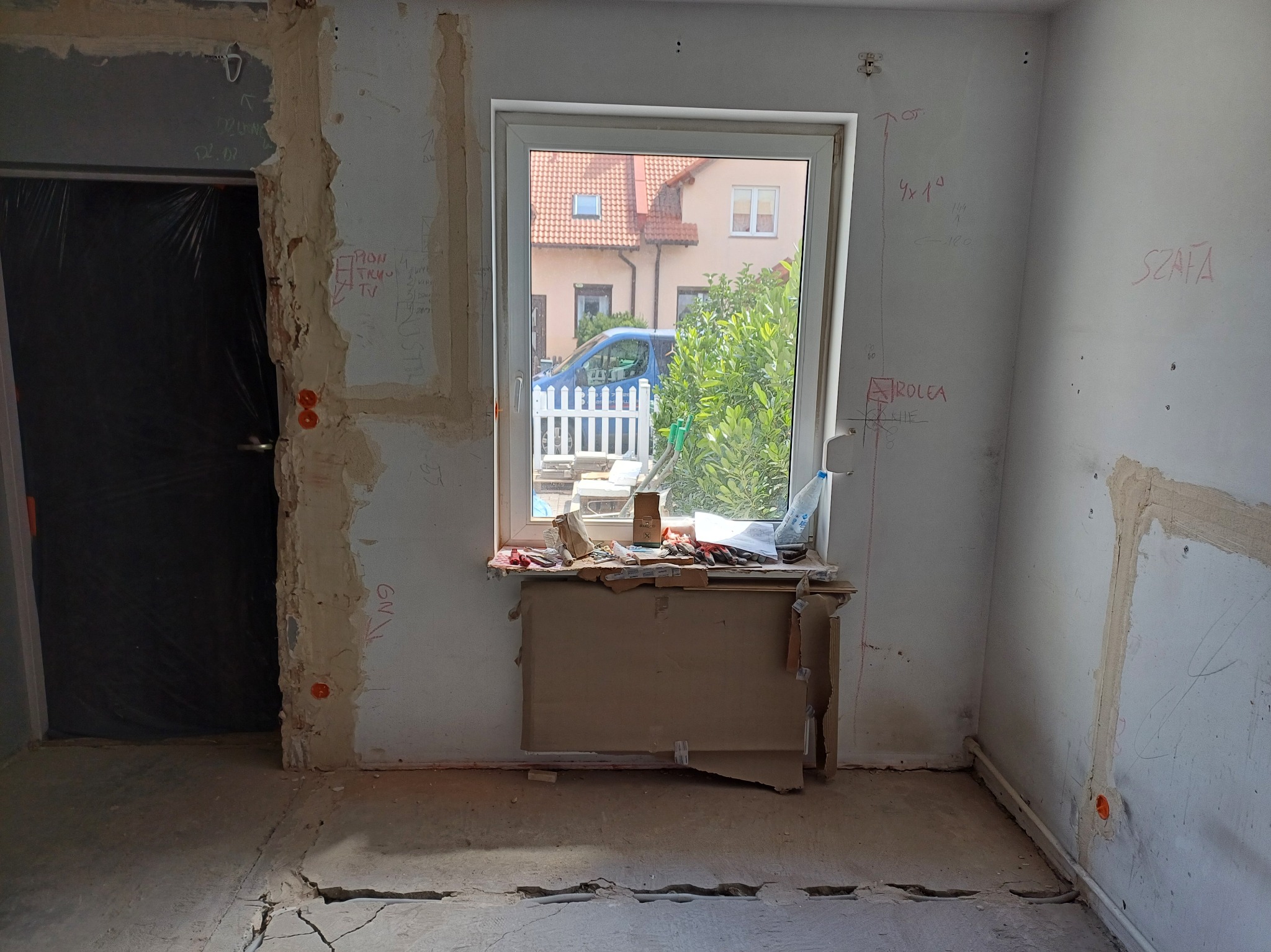 Wnętrze w trakcie remontu: odsłonięte ściany z instalacjami, okno z widokiem na dom, prowizoryczne zabezpieczenia. Widoczne ślady po instalacjach elektrycznych.
