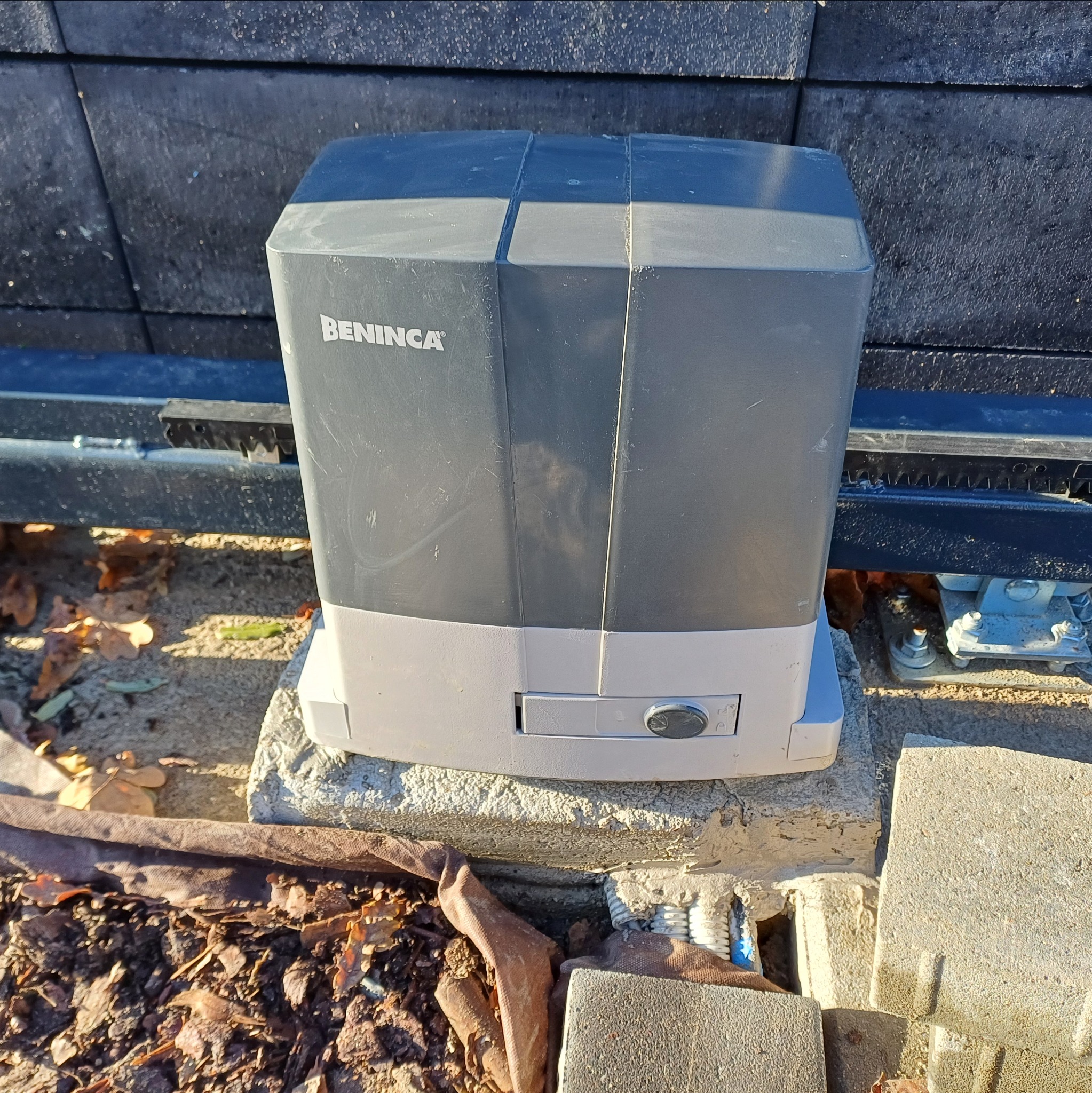 Szary napęd do bramy przesuwnej marki Beninca zamontowany na betonowej podstawie, widoczna listwa zębata i fragment ogrodzenia z betonowych bloczków.