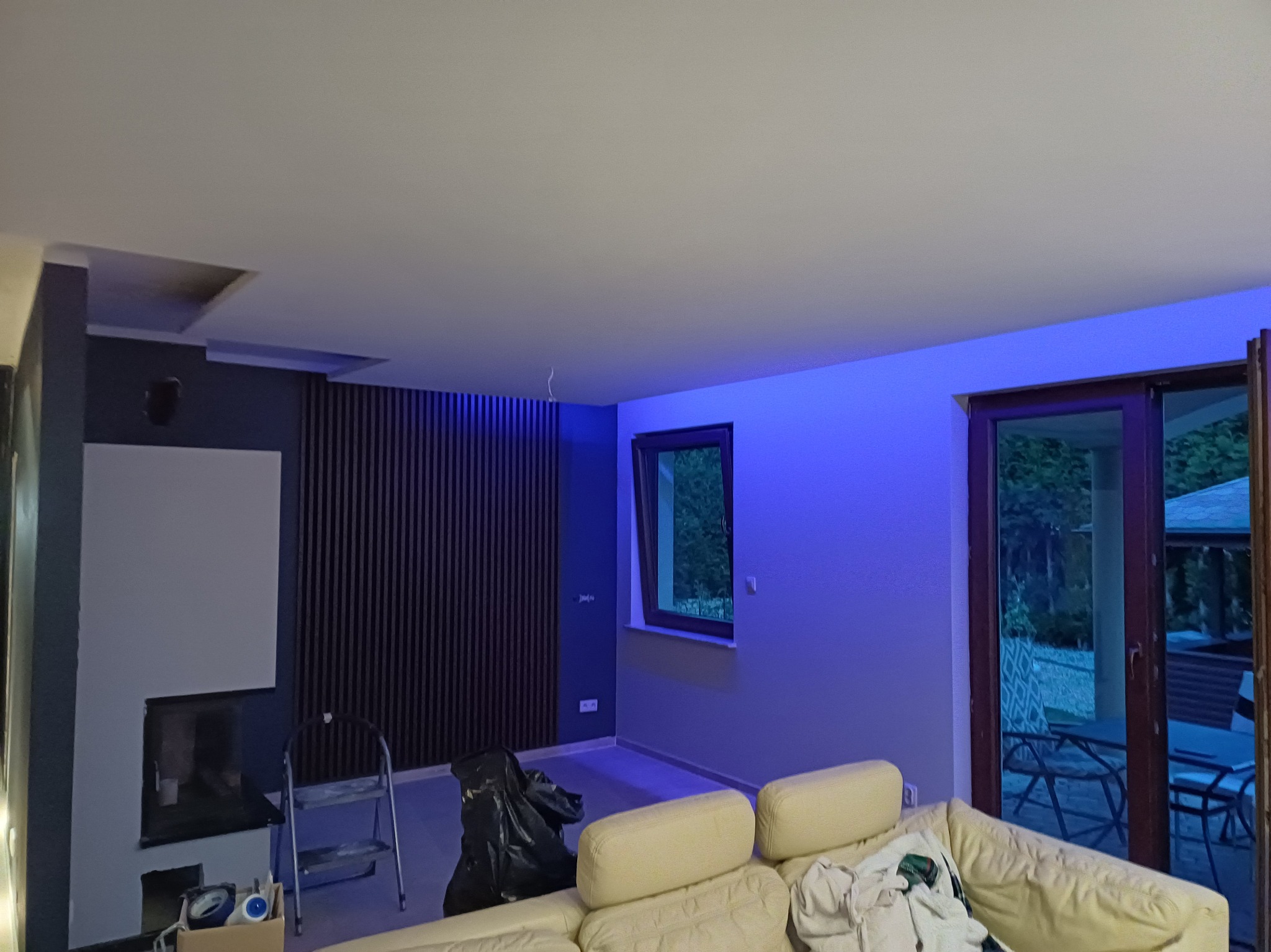 Wnętrze pokoju z kominkiem w trakcie prac wykończeniowych: widoczne elementy dekoracyjne, drabina malarska i worki z odpadami budowlanymi. Oświetlenie ambientowe.