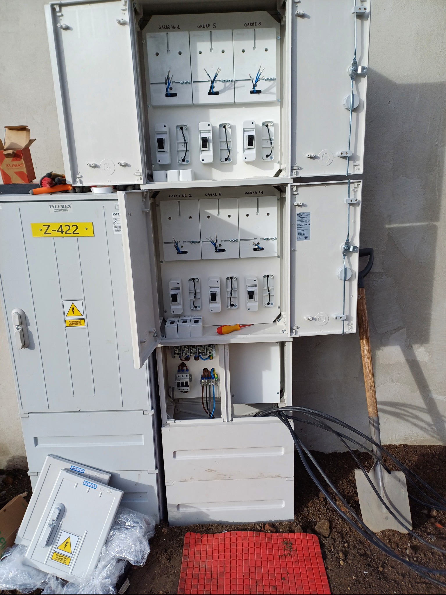 Otwarta, biała szafa elektryczna zewnętrzna z bezpiecznikami i przewodami, z żółtą tabliczką Z-422. Obok leżą pokrywy i narzędzia, a w tle widać szpadel.