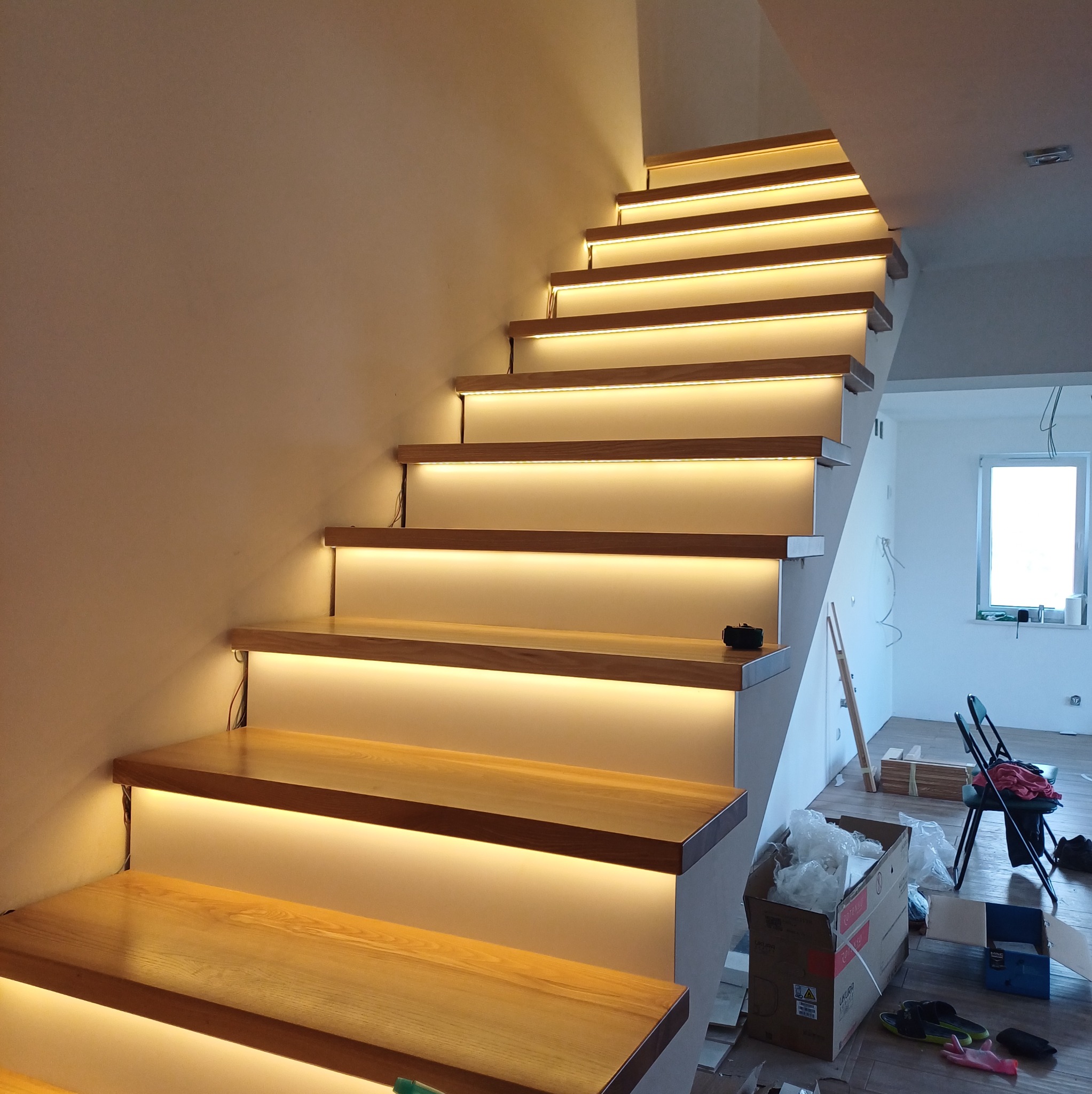 Schody z drewnianymi stopniami i subtelnym oświetleniem LED pod każdym stopniem, tworzącym ciepłą, nastrojową atmosferę. Widoczne okablowanie i elementy montażowe.