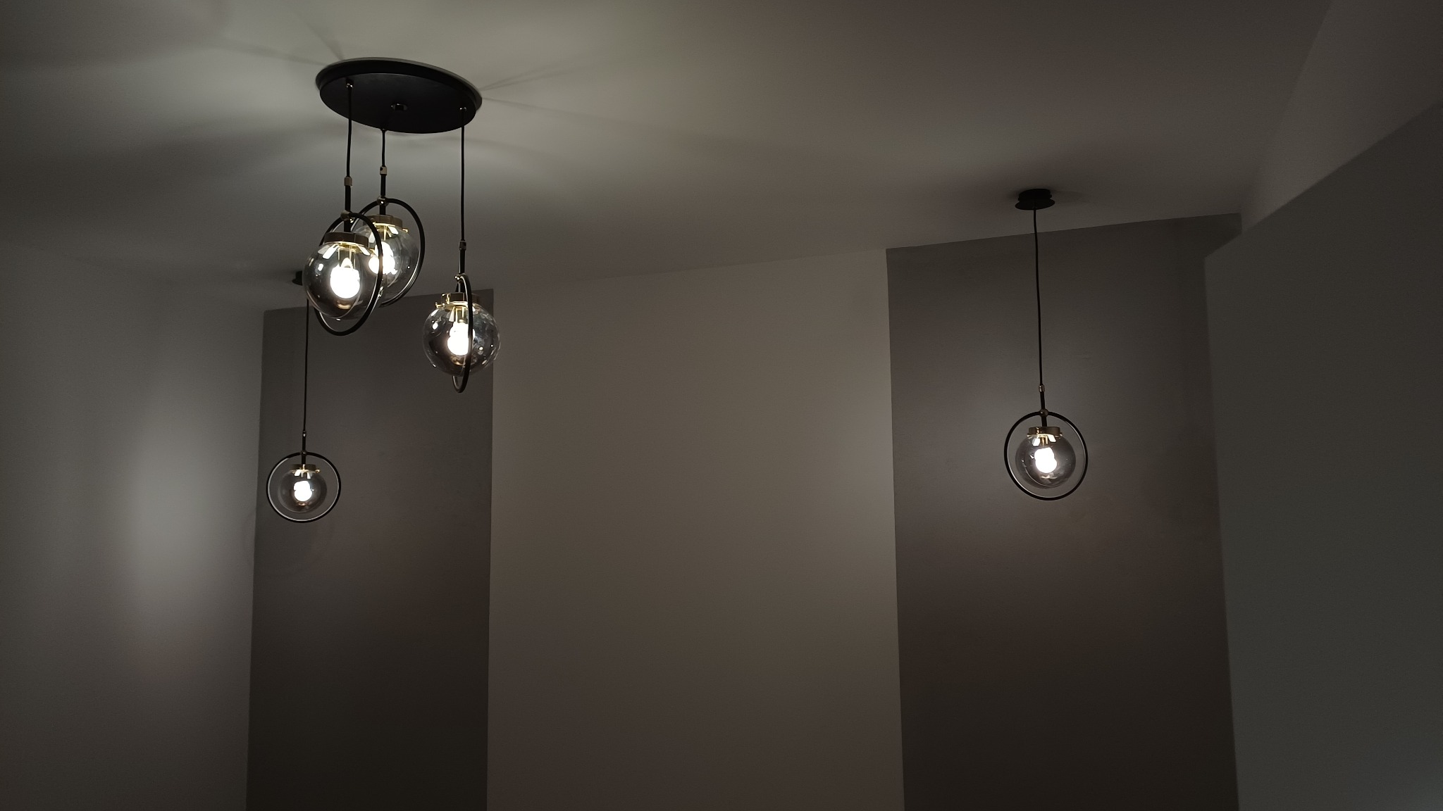 Nowoczesny żyrandol z trzema szklanymi kulami i pojedyncza lampa wisząca na tle szarej ściany. Minimalistyczny design oświetlenia w pomieszczeniu.