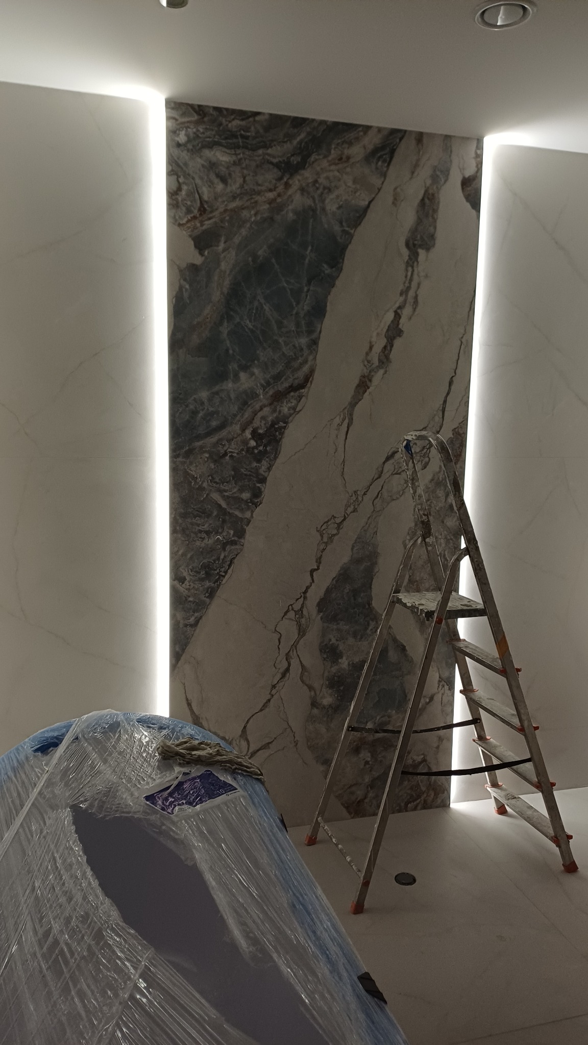 Montaż dekoracyjnej ściany z marmuru z podświetleniem LED. Widoczna drabina i zabezpieczone folią meble. Wnętrze w trakcie wykończenia.
