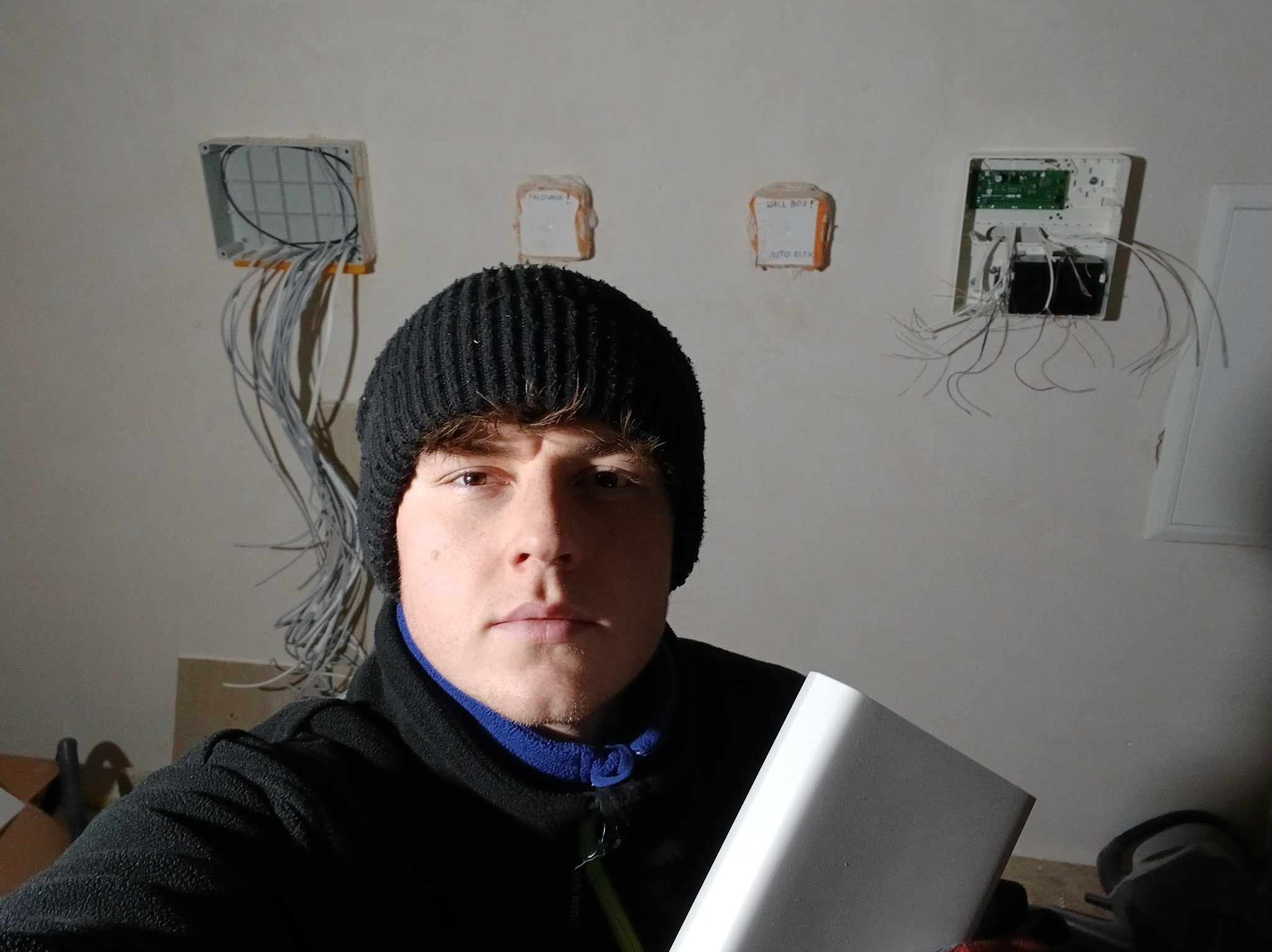 Mężczyzna w czapce na tle ściany z puszkami instalacyjnymi i przewodami. Widoczne elementy instalacji alarmowej/elektrycznej w trakcie montażu.