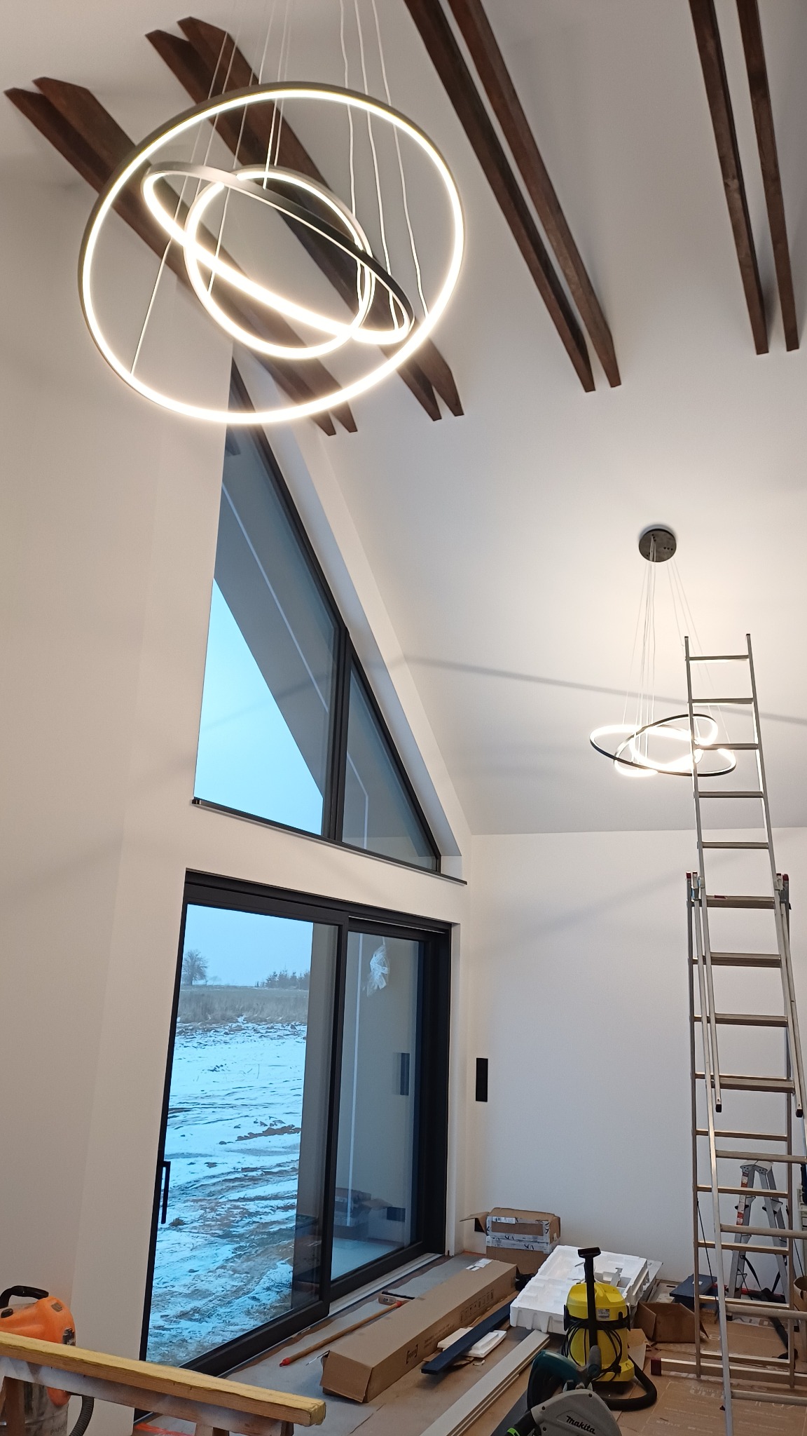 Nowoczesne, okrągłe lampy LED zamontowane w wysokim salonie z drewnianymi belkami stropowymi i dużym oknem. Widoczna drabina i narzędzia.