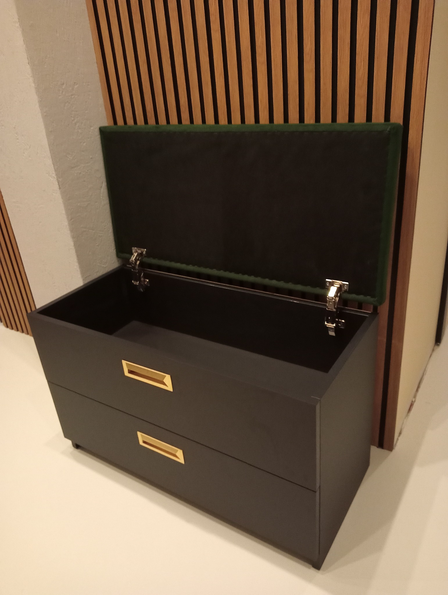 Stylowa, grafitowa komoda z dwoma szufladami i otwieranym od góry schowkiem z zielonym obiciem, widoczne złote uchwyty, tło z drewnianych paneli.