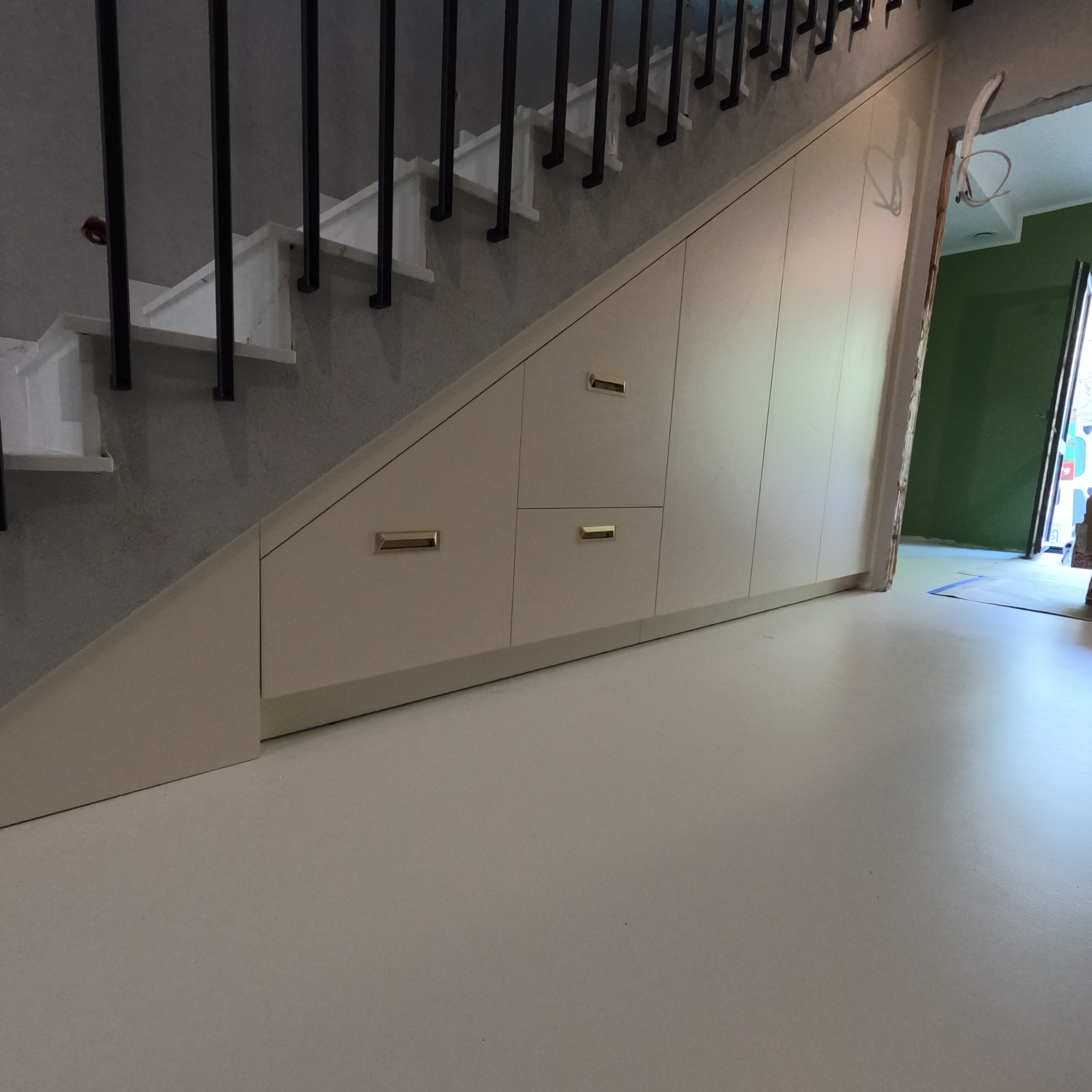Zabudowa schodów z szufladami i szafkami w kolorze kremowym, z minimalistycznymi złotymi uchwytami. Nowoczesne wykończenie wnętrza, widok z dołu na schody.