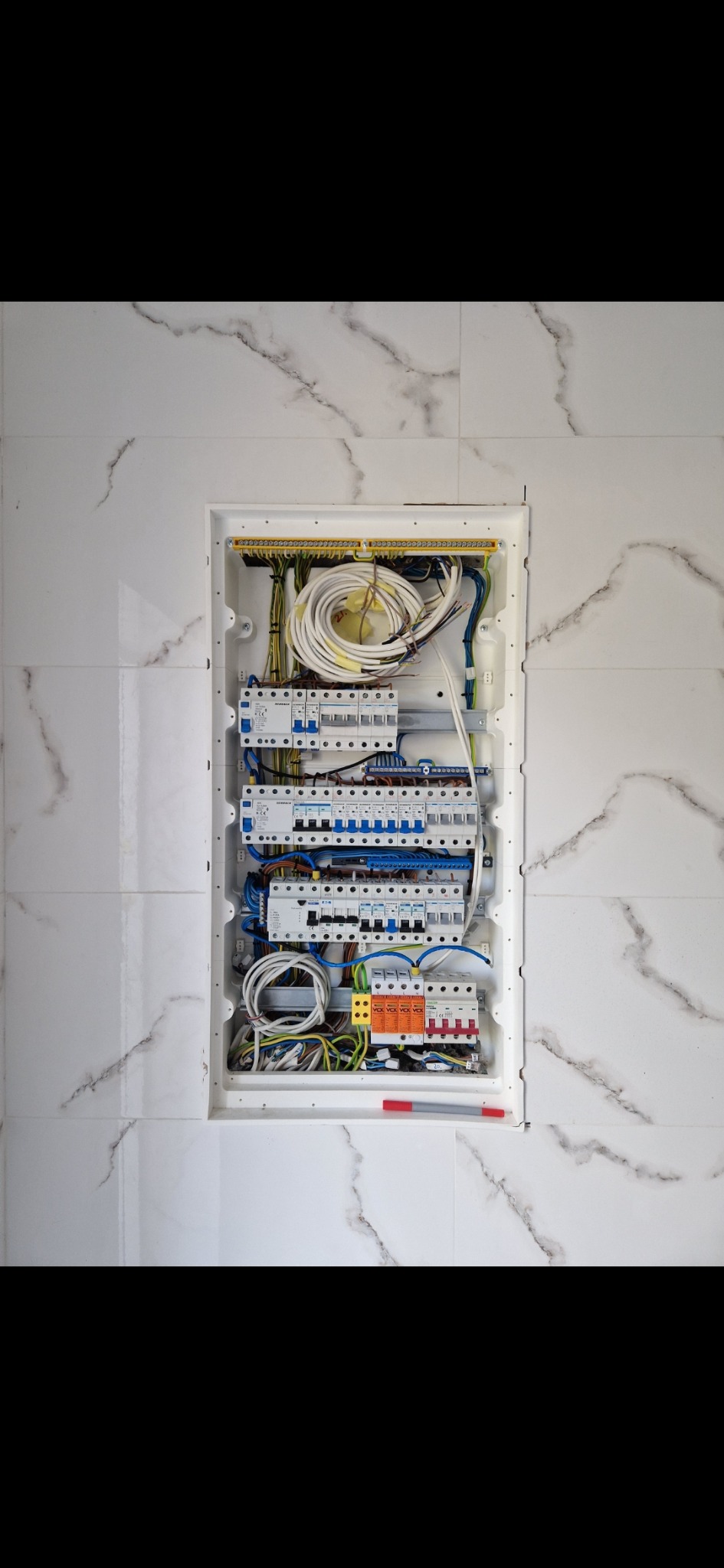 Rozdzielnica elektryczna z bezpiecznikami i przewodami, zamontowana w ścianie wyłożonej białymi płytkami imitującymi marmur.