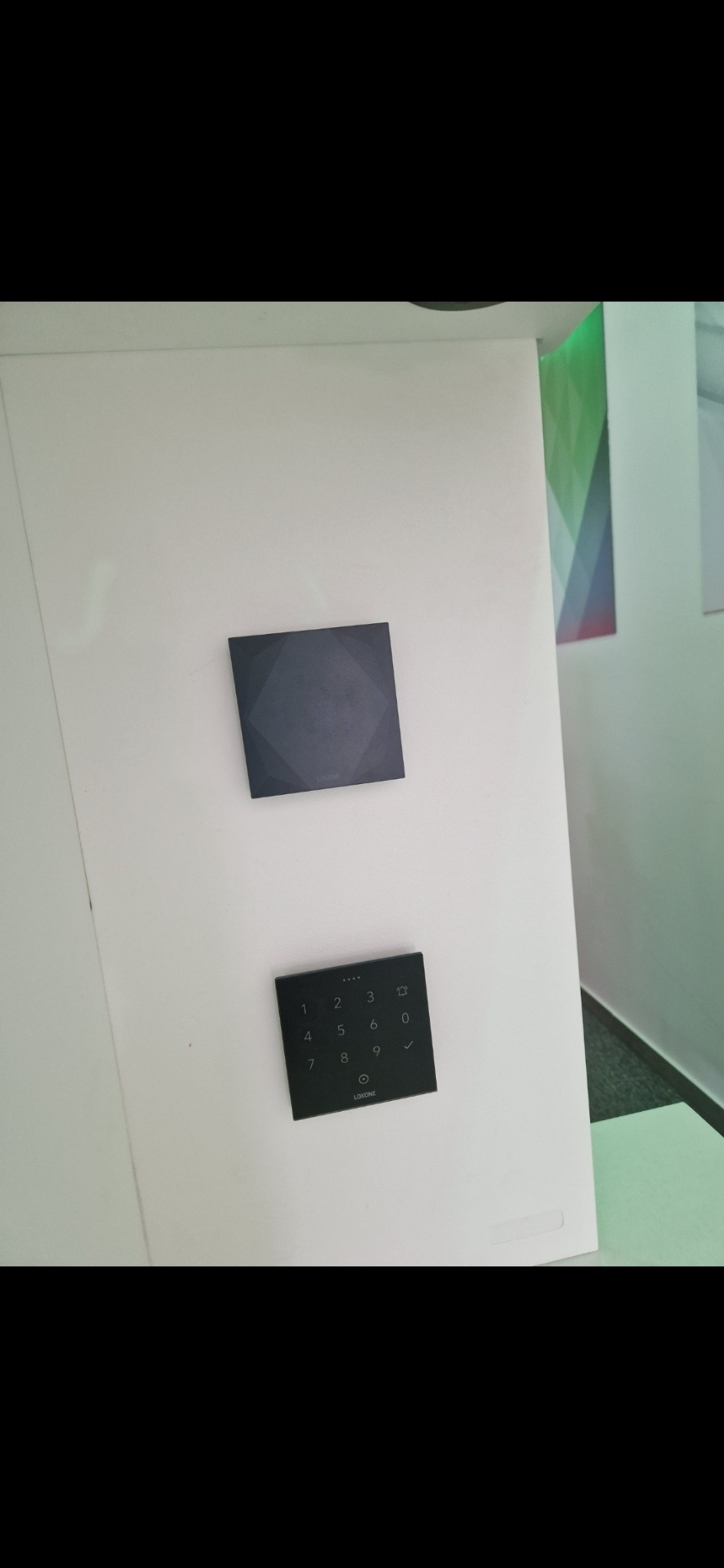 Dwa czarne panele Loxone na białej ścianie, jeden z klawiaturą numeryczną, drugi gładki z subtelnym wzorem. Nowoczesny design, minimalistyczny styl.