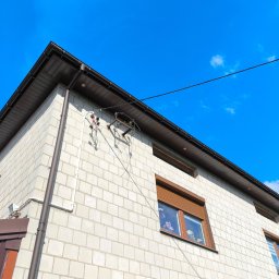 ElektrWit - Narożnik domu z białej cegły z widoczną instalacją elektryczną na ścianie i brązowym orynnowaniem na tle błękitnego nieba.