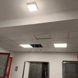 ElektrWit - Sufit podwieszany z otwartym panelem, widoczne przewody. W tle osoba w windzie. Oświetlenie sufitowe. Prace remontowe lub naprawa. Widok z dołu.