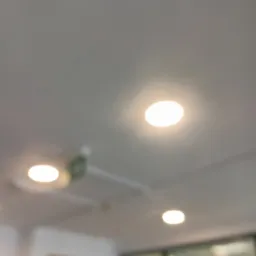 Biały sufit z trzema okrągłymi, wpuszczanymi lampami LED. Jedna z lamp częściowo zasłonięta przez element wentylacyjny. Rozproszone światło.