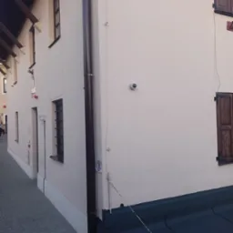 Elewacja budynku z kamerą monitoringu, brązową rynną i drewnianymi okiennicami. Widoczne kable i przewody na ścianie budynku.